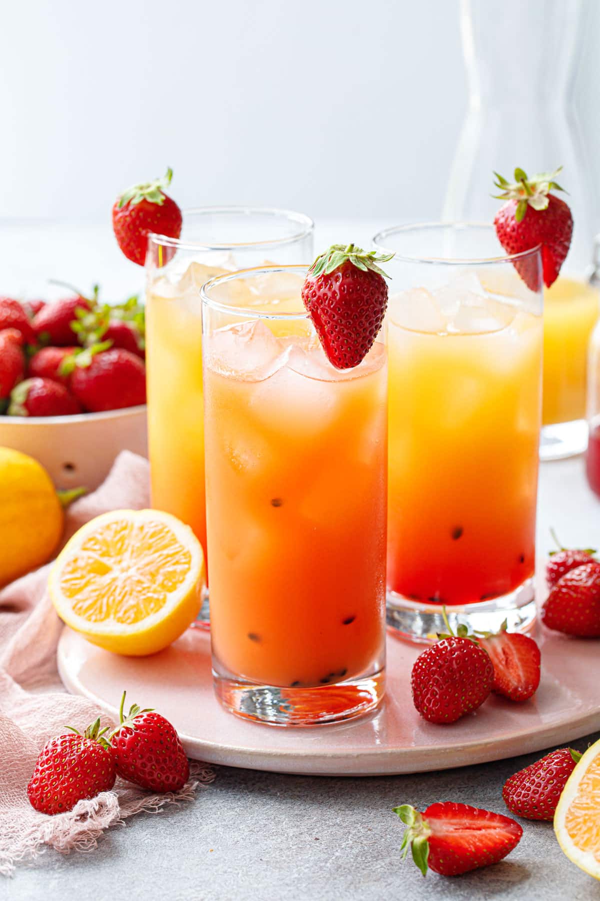 Three glasses of Strawberry Passionfruit Lemonade, one stirred for a solid orange color, the others showing a gradient red to yellow, with strawberries on the rim of each glass.