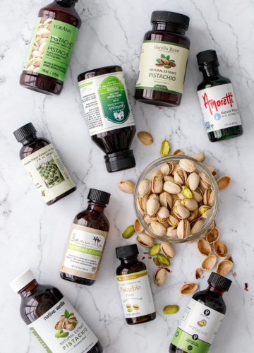 Nine bottles of different brands of pistachio extract, flat lay on a marble background with a bowl of pistachios.