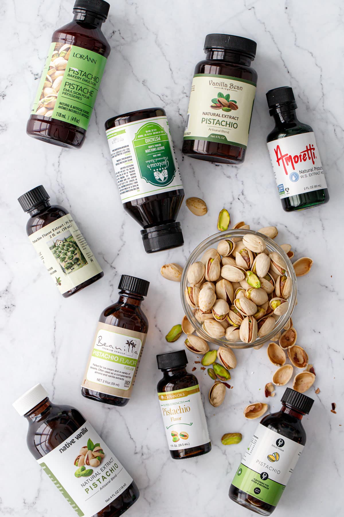 Nine bottles of different brands of pistachio extract, flat lay on a marble background with a bowl of pistachios.