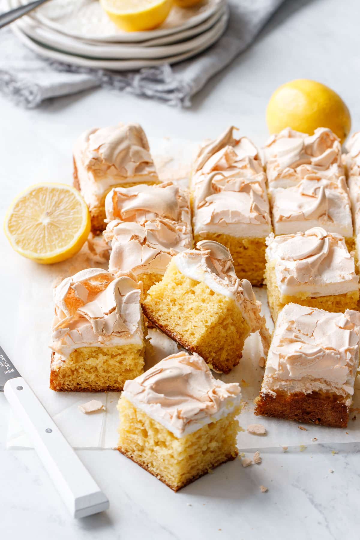 Cut squares of Lemon Meringue Butter Cake on a marble background with white knife and stack of plates in the background.