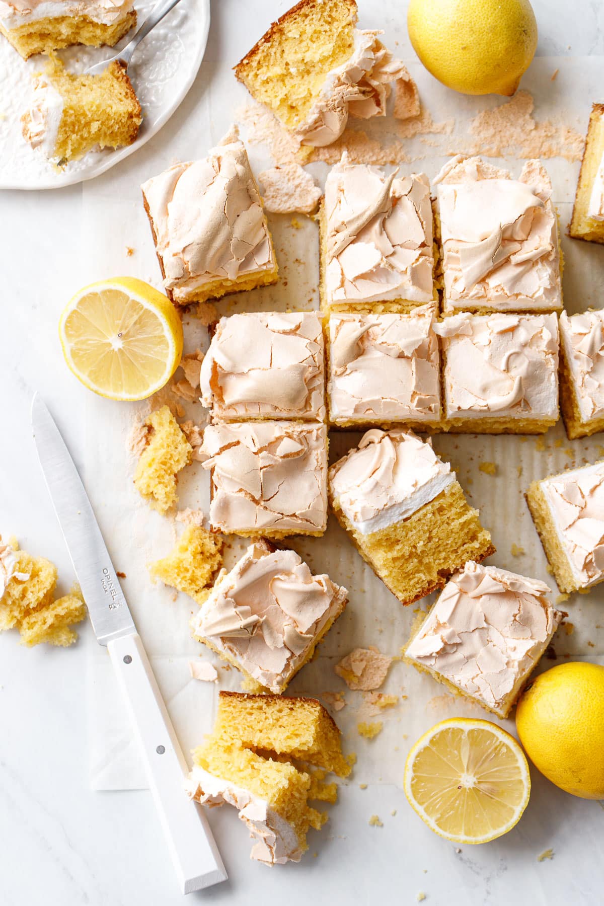 Overhead, messy pieces of Lemon Meringue Butter Cake cut into squares, with lemons (whole and cut in half) and a small knife.