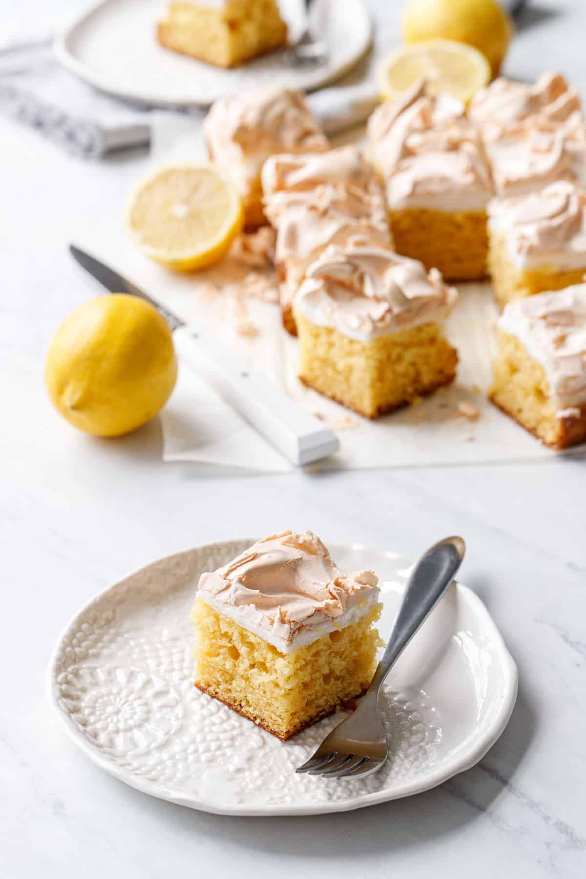 Square of Lemon Meringue Butter Cake on a porcelain plate with a fork, showing the moist crumb and toasted meringue layer on top.