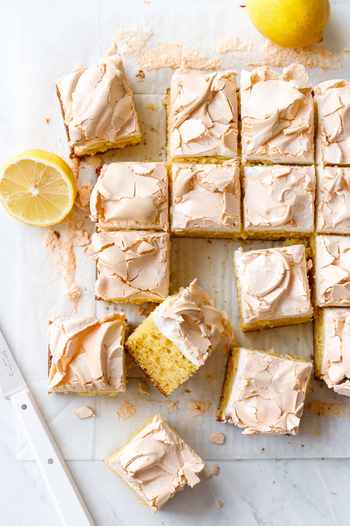 Overhead, cut squares of Lemon Meringue Butter Cake with knife and cut lemons on marble and parchment paper.