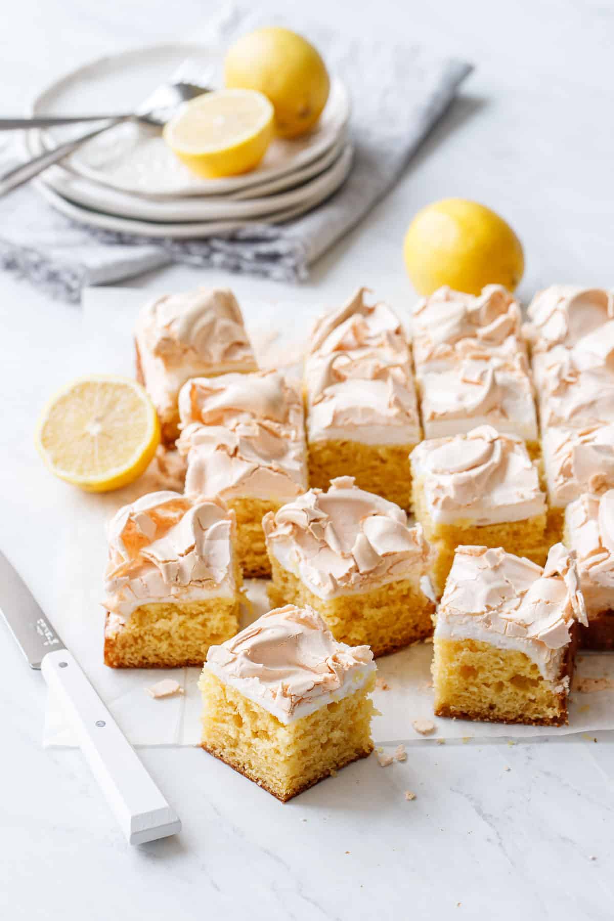 Lemon Meringue Butter Cake cut into squares on a piece of parchment on marble, with a knife, lemons and stack of plates in the background.