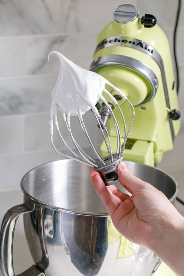 Hand holding wire whisk attachment with meringue showing stiff peaks, green mixer in the background.