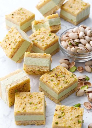 Cut squares of Pistachio Blondie Ice Cream Sandwiches scattered on a marble background with a bowl of pistachios on the side.