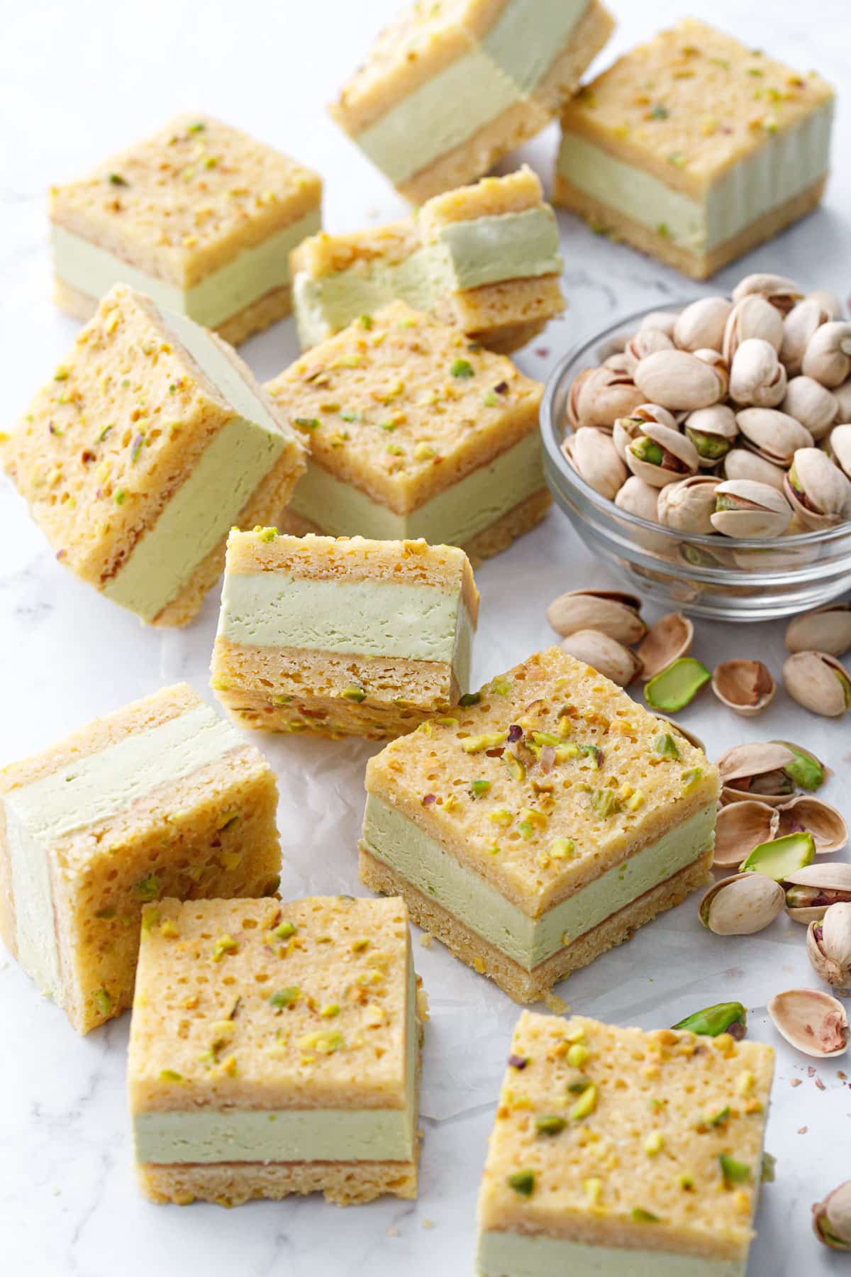 Cut squares of Pistachio Blondie Ice Cream Sandwiches scattered on a marble background with a bowl of pistachios on the side.