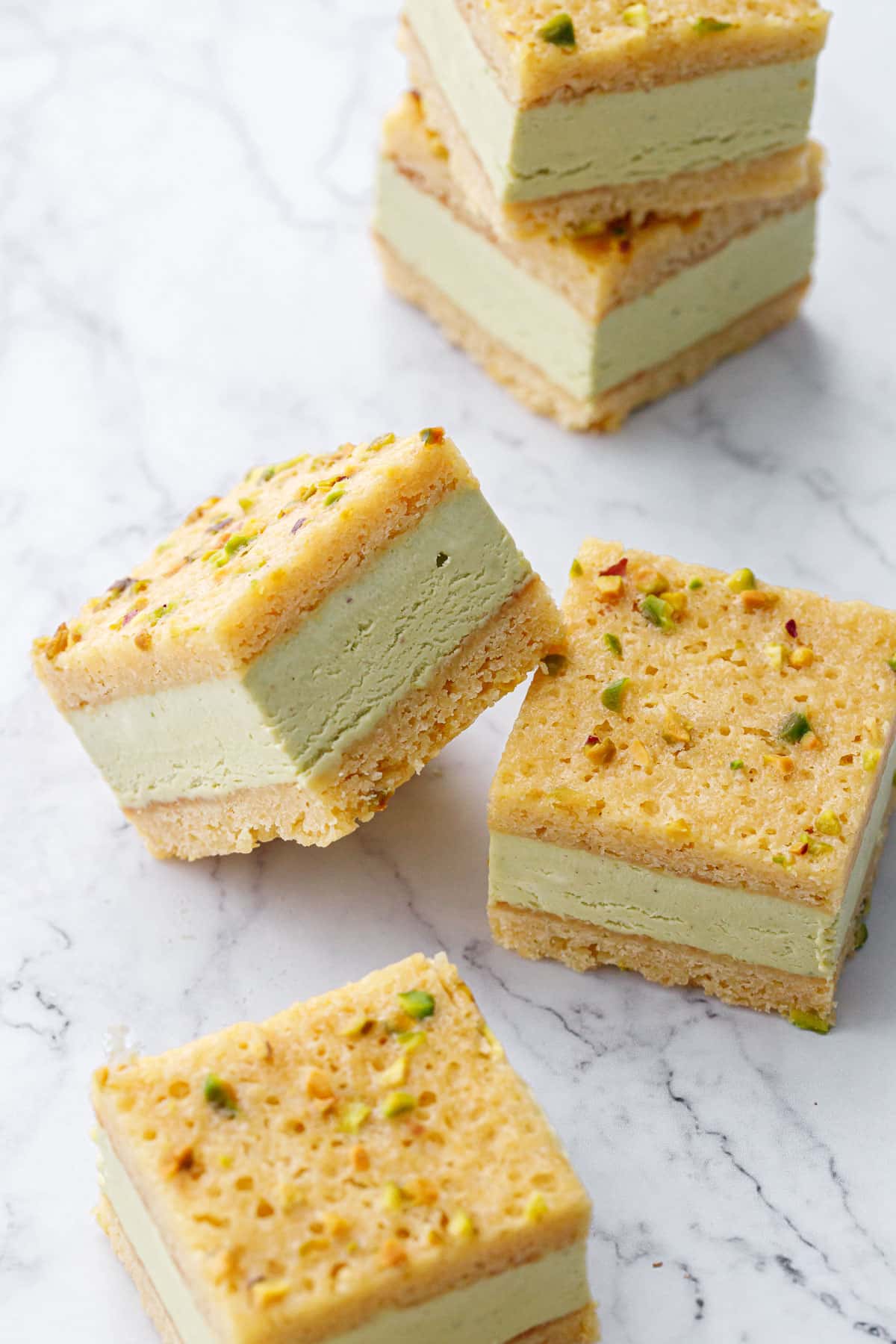 Perfectly cut squares of Pistachio Blondie Ice Cream Sandwiches on a marble background, showing the clear layers of blondie and creamy no-churn pistachio ice cream sandwiched in between.