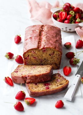 Loaf of Strawberry Banana Bread with a pink glaze, two slices laying down to show the interior texture, and strainer with strawberries in the background.