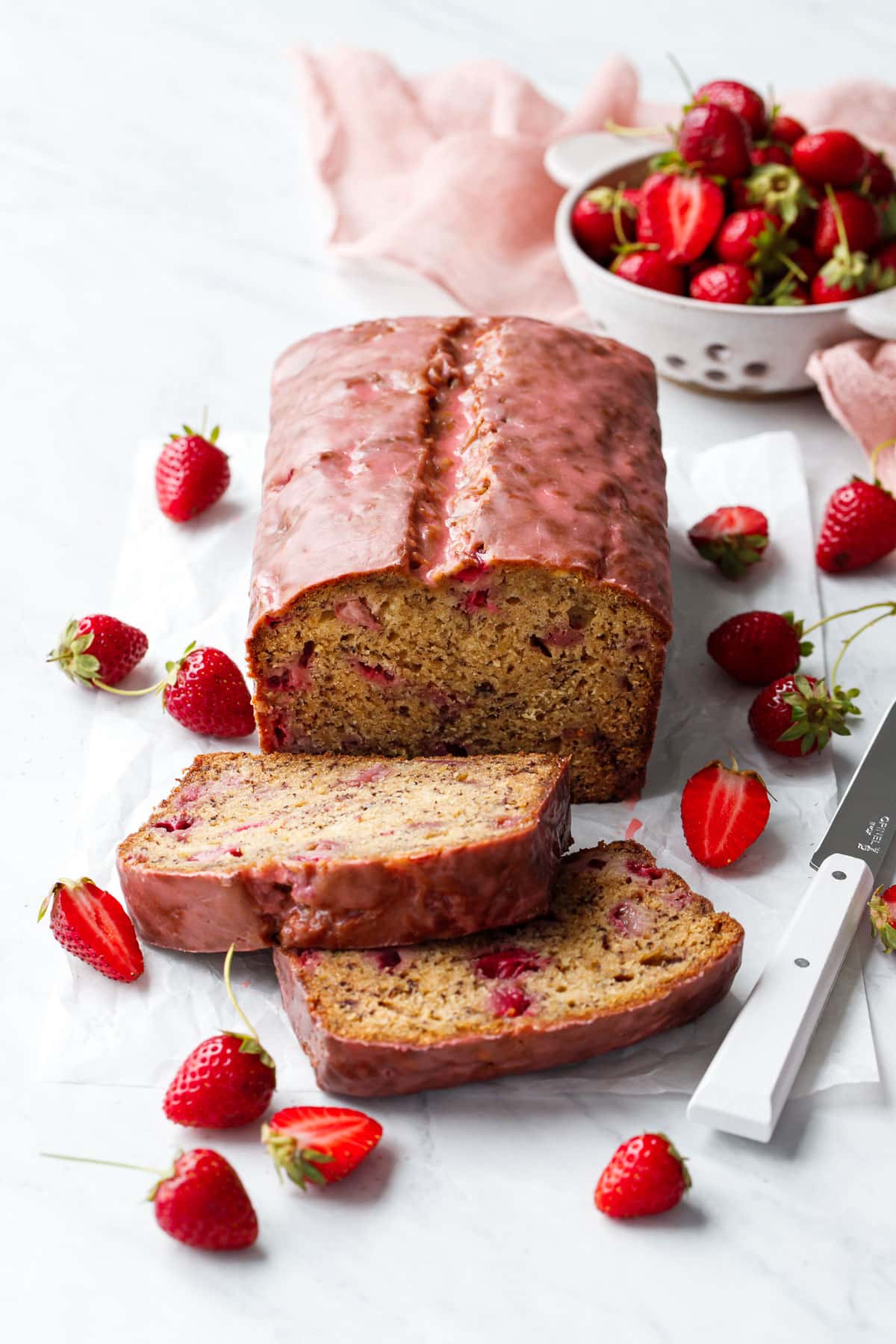 Loaf of Strawberry Banana Bread with a pink glaze, two slices laying down to show the interior texture, and strainer with strawberries in the background.