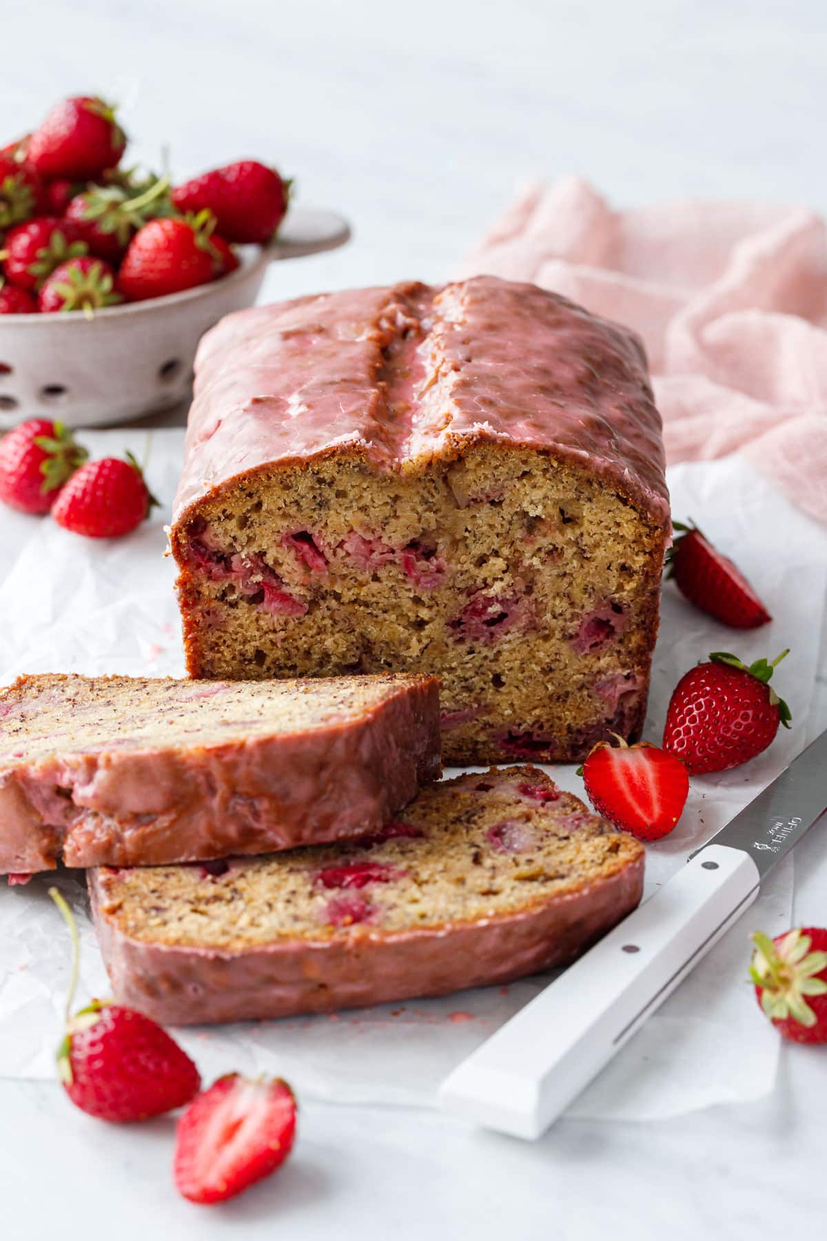 Loaf of Strawberry Banana Bread with a pink glaze, two slices laying down to show the interior texture, and strainer with strawberries in the background.