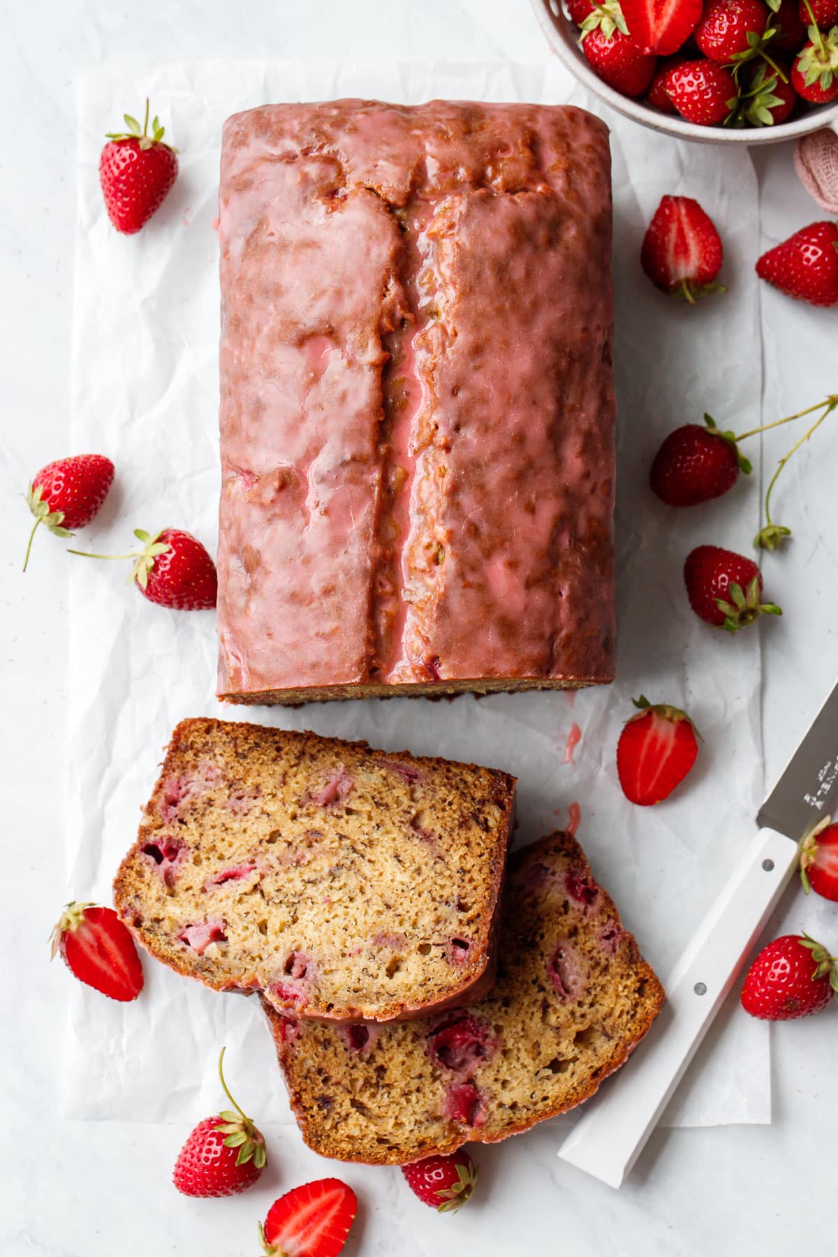 Overhead, loaf of Strawberry Banana Bread with two slices cut and laying down to show texture, scattered strawberries and a white knife on the side.