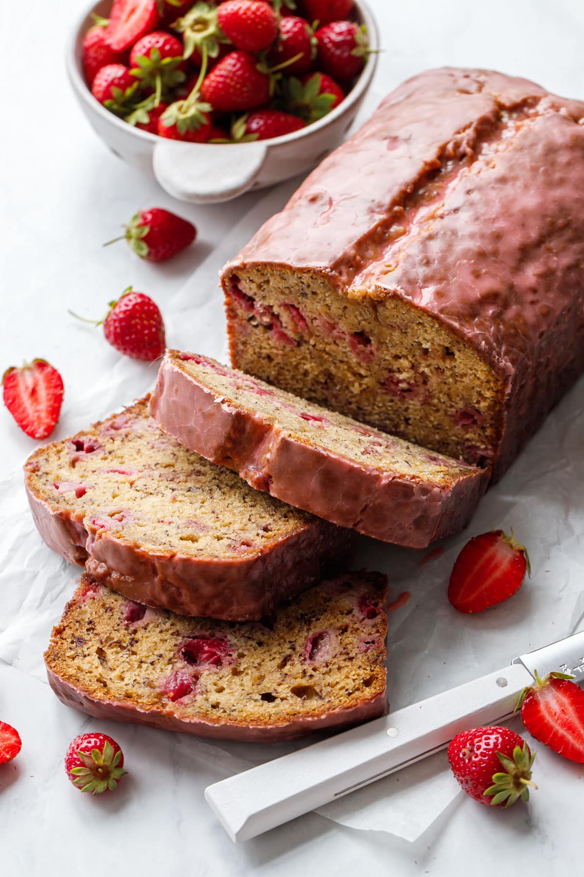 Loaf of Strawberry Banana Bread with a pink strawberry glaze, slices laying down to show the moist inner texture studded with pockets of fresh strawberries.