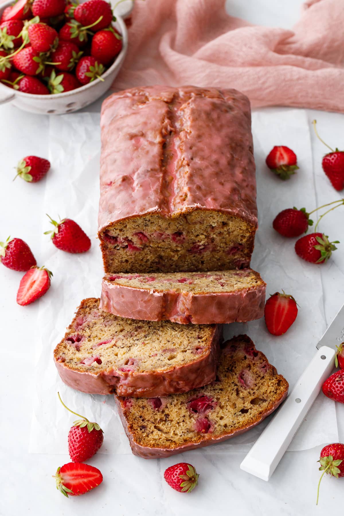 Loaf of Strawberry Banana Bread with a pink glaze, two slices laying down to show the interior texture, and strainer with strawberries in the background.