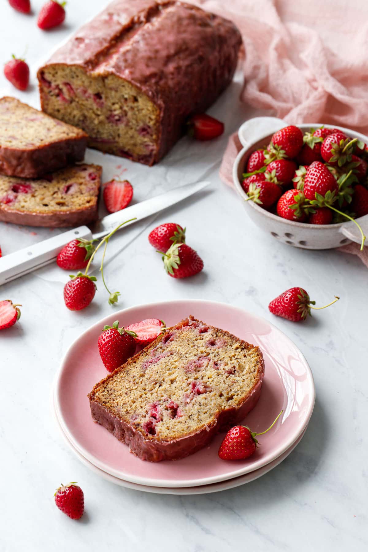 Slice of Strawberry Banana Bread on a pink plate, with the full loaf and slices in the background with a bowl of fresh strawberries and a knife.
