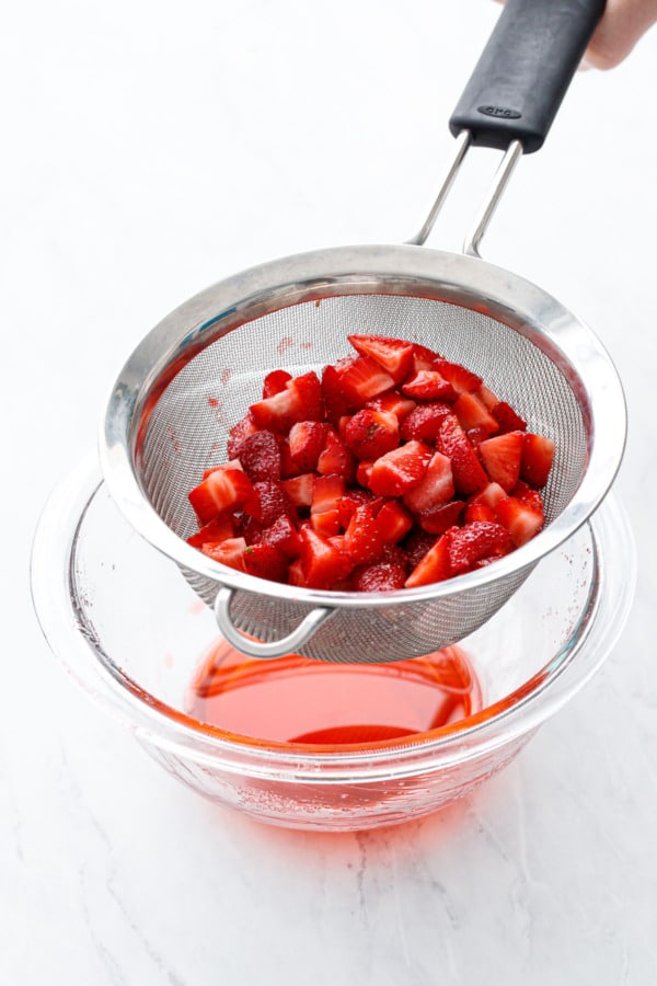 Straining fresh strawberry pieces in a fine mesh sieve, liquid collected in a glass bowl below.