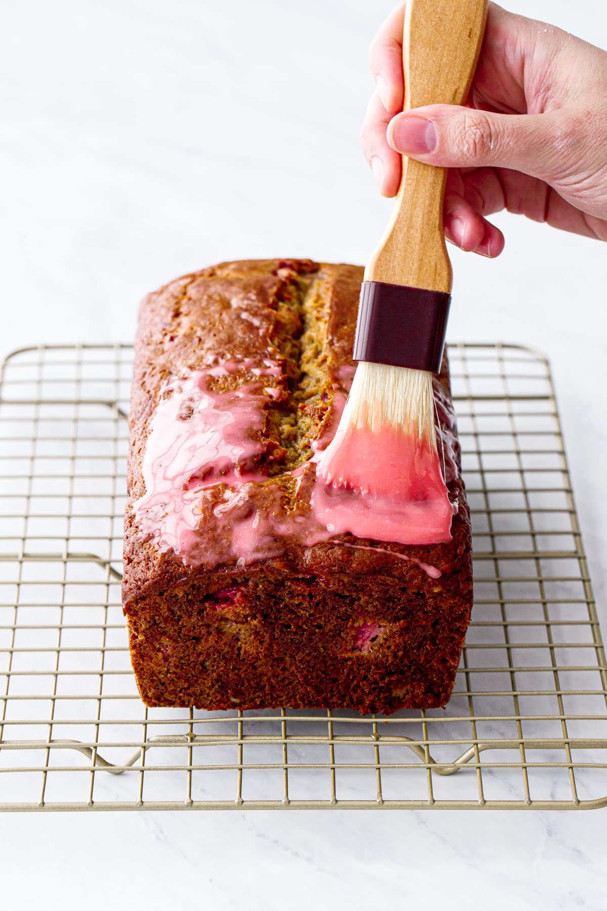 Brushing the baked loaf of banana bread with a bright pink strawberry glaze using a pastry brush.