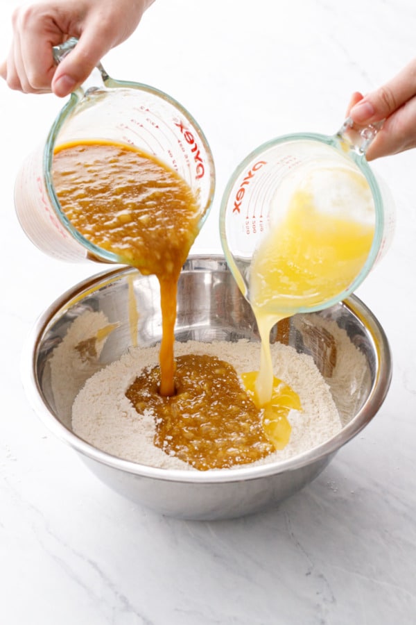Pouring the mashed banana mixture and melted butter into a metal mixing bowl with dry ingredients.
