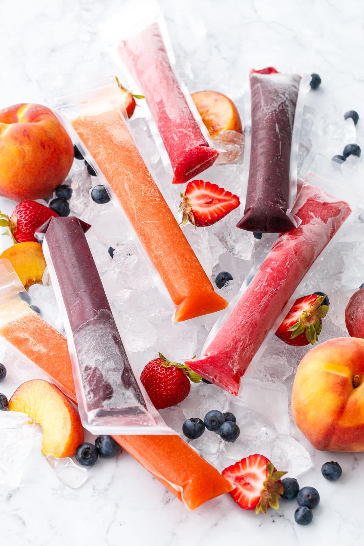 Freezer Jam Ice Pops in three different color/flavors, scattered on crushed ice cubes with fresh strawberries, blueberries, and peaches scattered around.