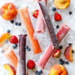 Overhead, Freezer Jam Ice Pops in three different color/flavors, scattered on crushed ice cubes with fresh strawberries, blueberries, and peaches scattered around.