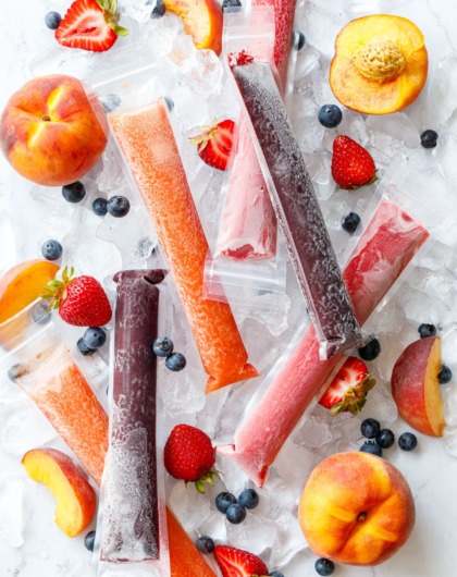 Overhead, Freezer Jam Ice Pops in three different color/flavors, scattered on crushed ice cubes with fresh strawberries, blueberries, and peaches scattered around.