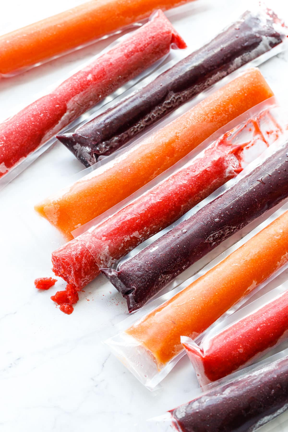 Rows of alternating colors/flavors of Freezer Jam Fruit Ice Pops, one strawberry popsicle cut and with a bite out of it to show the texture.