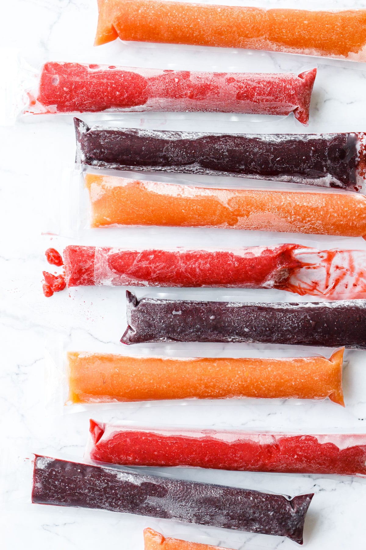 Overhead, row of alternating colors of peach, blueberry, and strawberry freezer jam ice pops, one with a bite out of it to show texture.