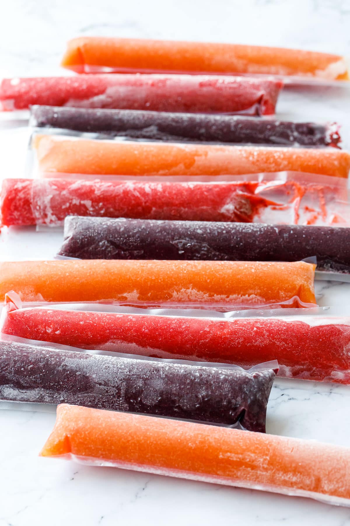 Row of alternating colors of peach, strawberry, and blueberry ice pops with frost and condensation, on a marble background.