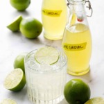 Bottles of Homemade Lime Cordial with fresh limes and a gimlet cocktail garnished with a lime slice.