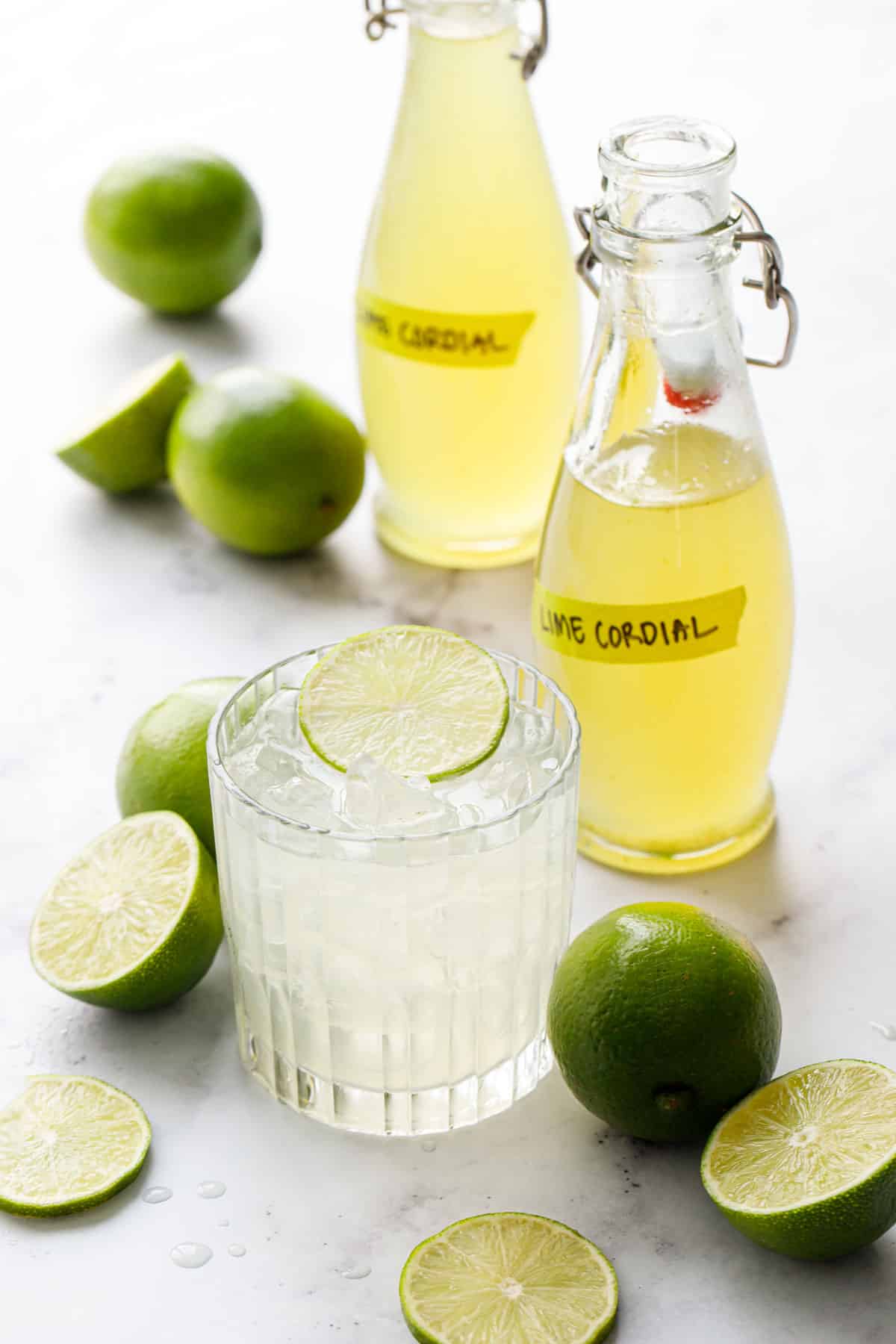 Bottles of Homemade Lime Cordial with fresh limes and a gimlet cocktail garnished with a lime slice.