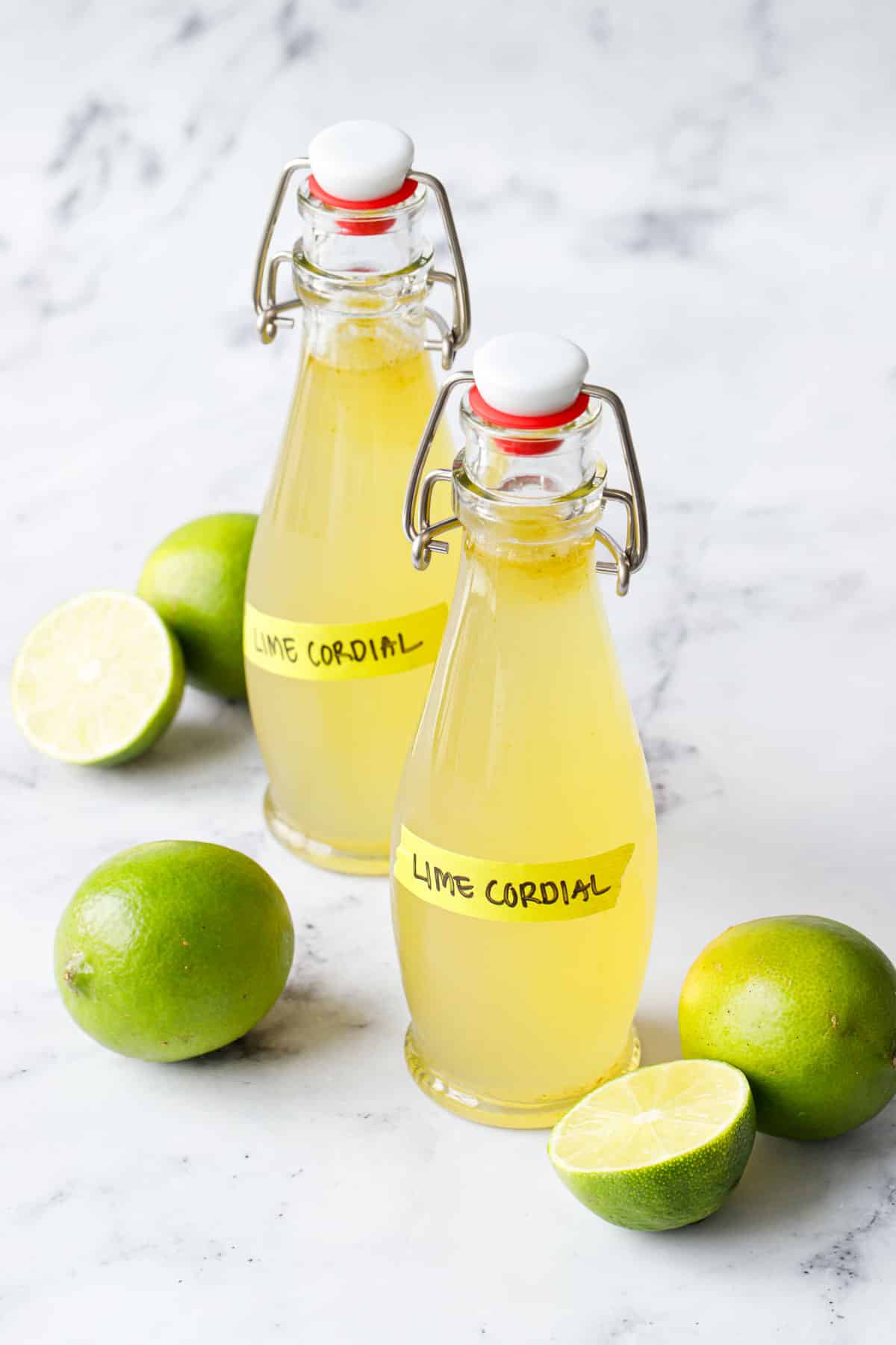 Two swingtop bottles filled with Homemade Lime Cordial on a marble background with fresh limes.