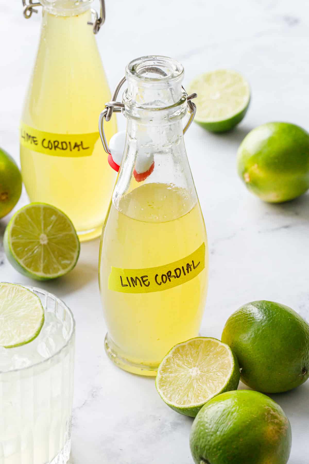 Swing-top bottles filled with Homemade Lime Cordial, labeled with green tape, with fresh limes scattered around.