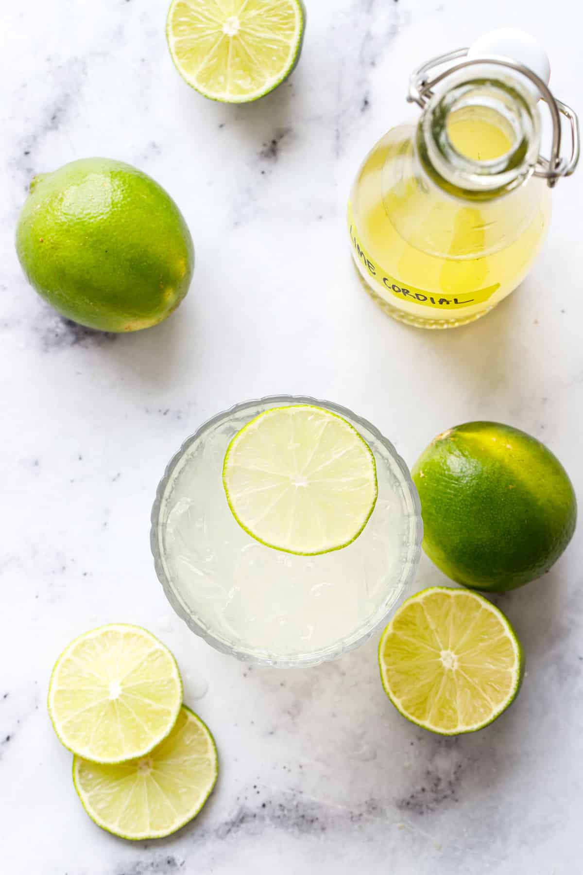 Overhead, glass with a lime Gimlet cocktail garnished with a lime slice, plus fresh limes and a bottle of Homemade Lime Cordial.