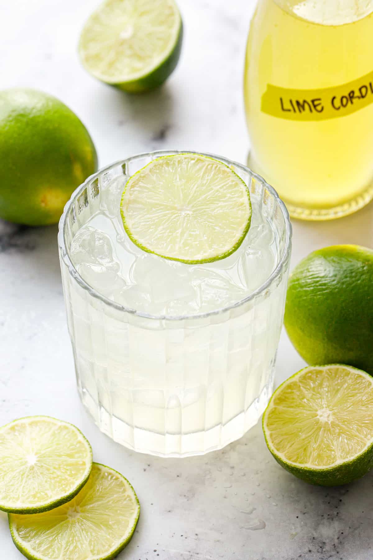 Gimlet cocktail in a lowball glass garnished with a lime slice, with bottle of Homemade Lime Cordial and fresh limes in the background.