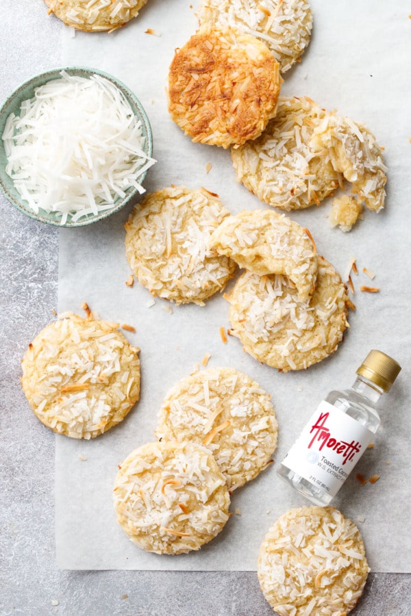 Overhead, scattered Toasted Coconut Sugar Cookies on parchment paper, with a bowl of coconut and a bottle of coconut extract off to the side.
