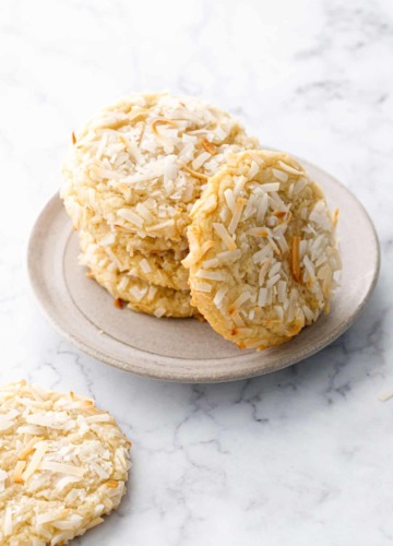 Stack of Toasted Coconut Sugar Cookies on a plate with one cookie leaning up against the stack to show the toasted shredded coconut on top of the cookie.