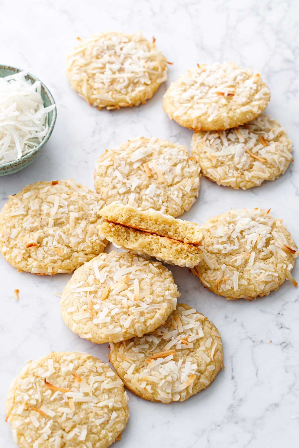 Messy arrangement of Toasted Coconut Sugar Cookies on a marble background, one cookie broken in half to show the inside texture.
