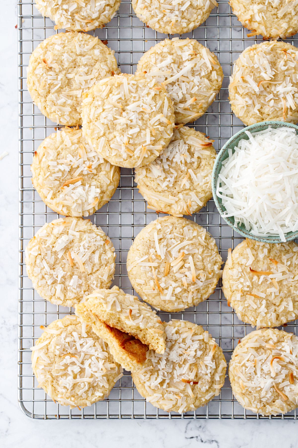 Overhead, rows of Toasted Coconut Sugar Cookies on a wire cooling rack, one cookie broken in half to show interior texture, plus a bowl of shredded coconut on the side.