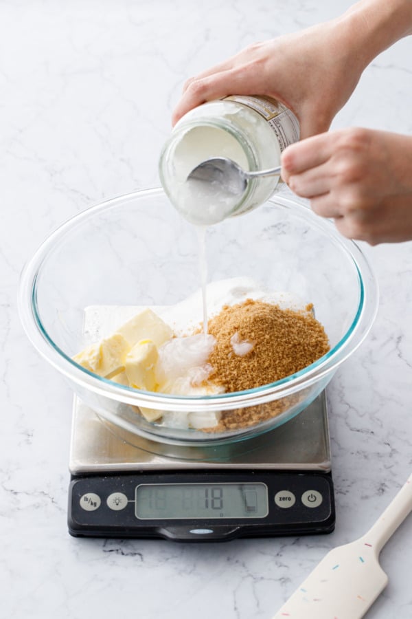 Glass mixing bowl on a scale, pouring coconut oil into bowl with other ingredients.