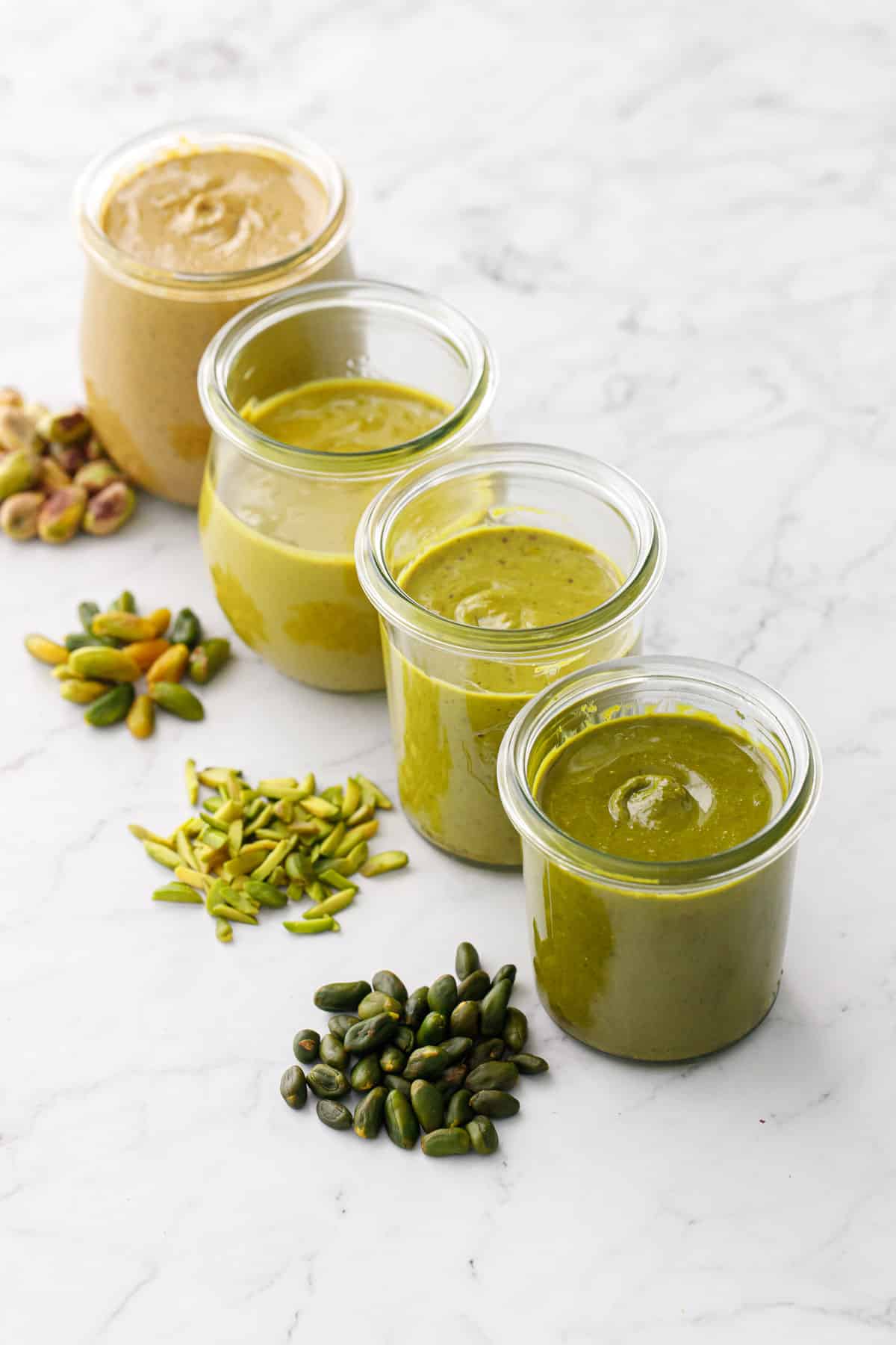 Four jars of Homemade Pistachio Butter in varying colors grading from brown to green, with the nut used to make it in a pile next to each jar.
