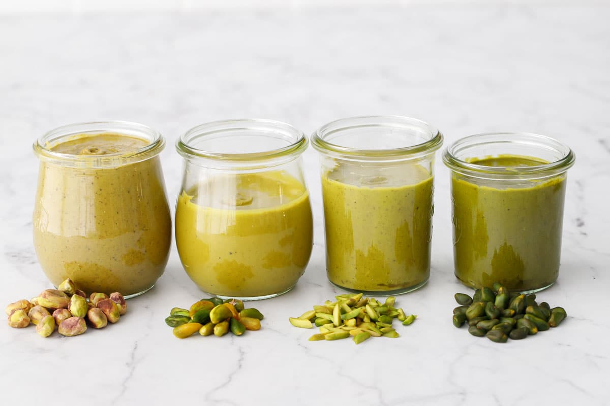 Four jars of Homemade Pistachio Butter in varying colors grading from brown to green, with the nut used to make it in a pile in front of each jar.