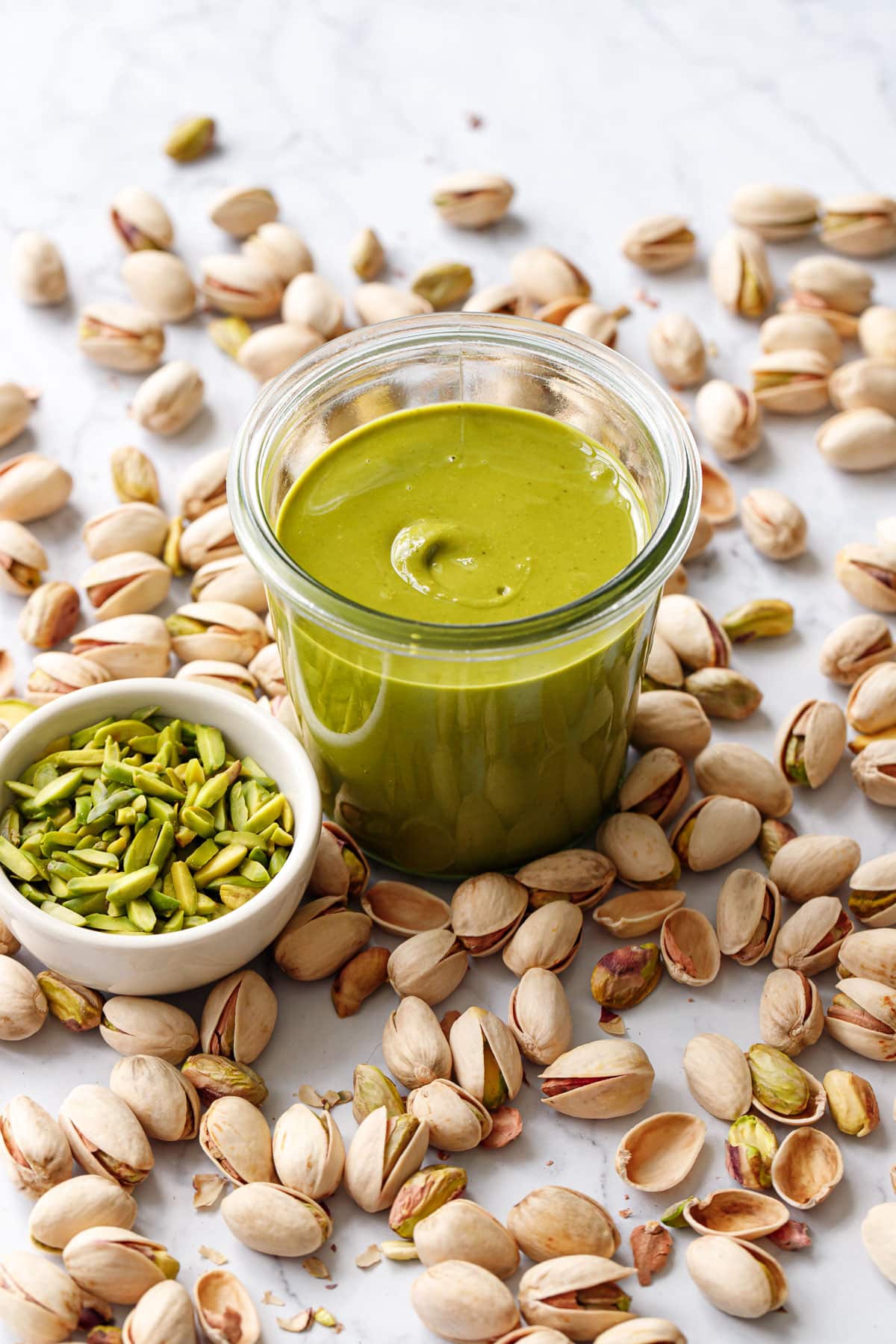 Glass jar of bright green Homemade Pistachio Butter with a bowl of green slivered pistachios, and whole in-shell pistachios scattered around.