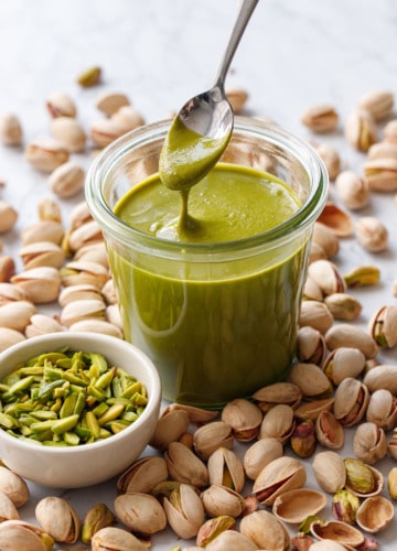 Glass jar with bright green Homemade Pistachio Butter, a spoonful lifting to show the creamy texture, with slivered and shell pistachios scattered around.