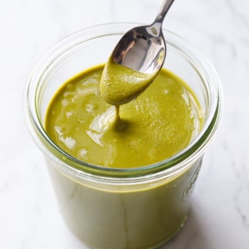 Glass jar of bright green Homemade Pistachio Butter on a marble background, spoonful lifted above to show the creamy texture.