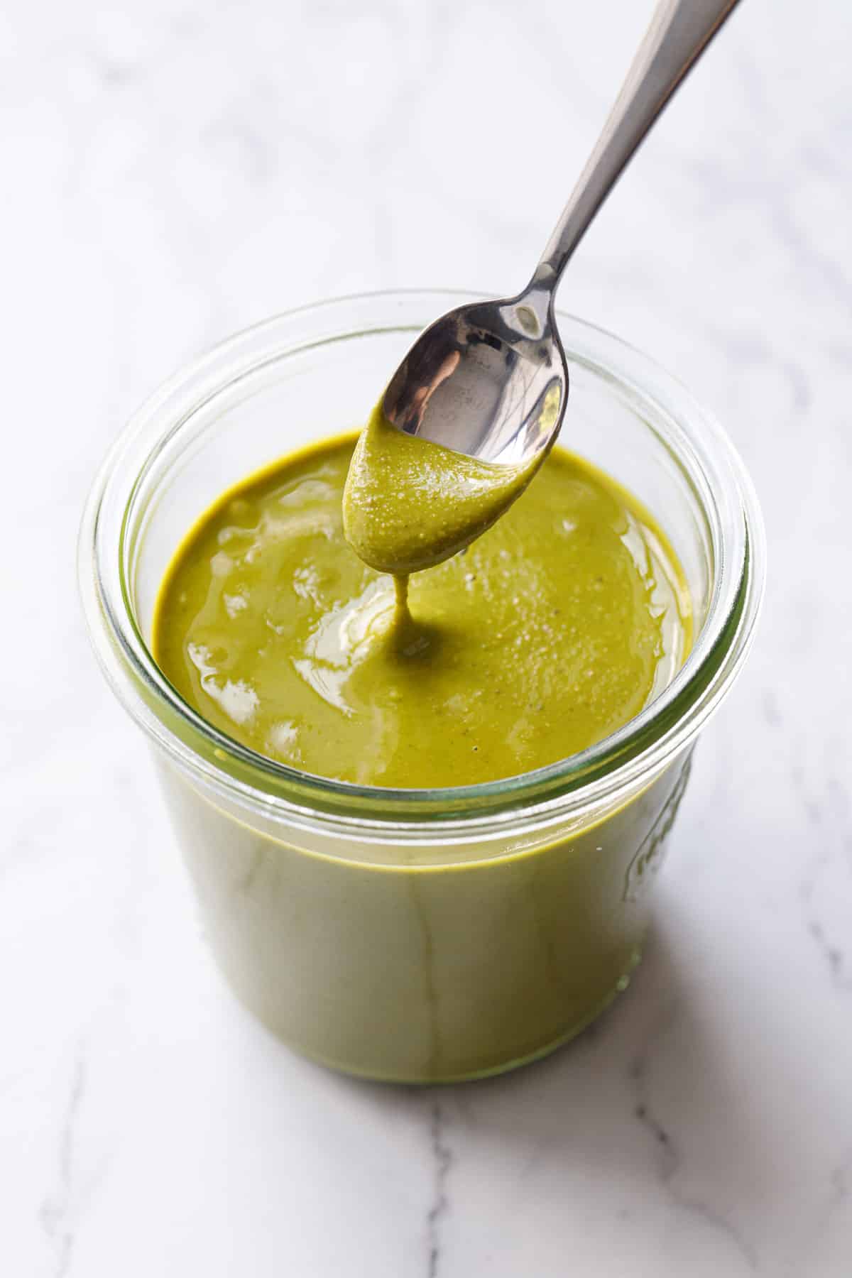 Glass jar of bright green Homemade Pistachio Butter on a marble background, spoonful lifted above to show the creamy texture.
