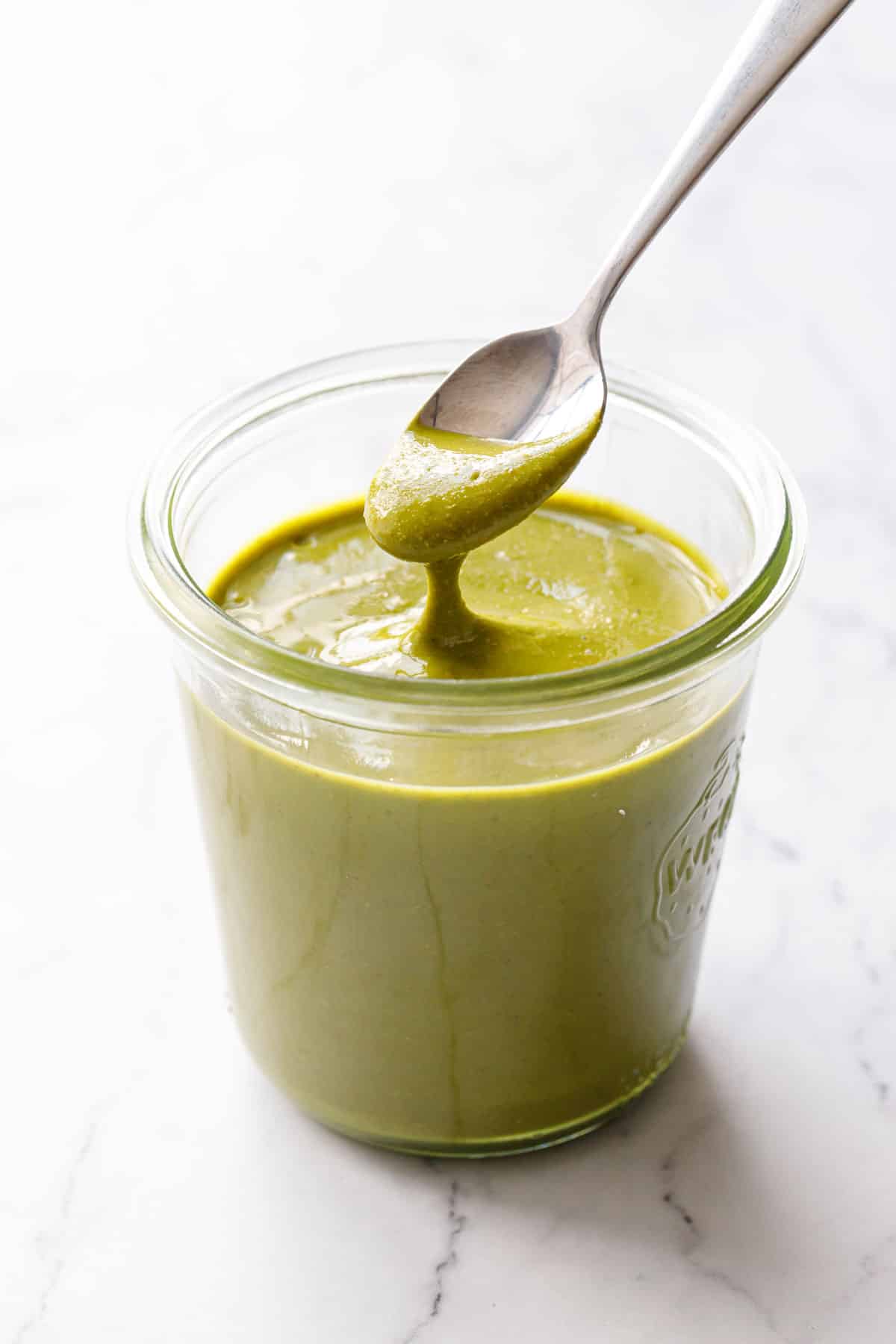 Glass jar of bright green Homemade Pistachio Butter on a marble background, spoonful lifted above to show the creamy texture.