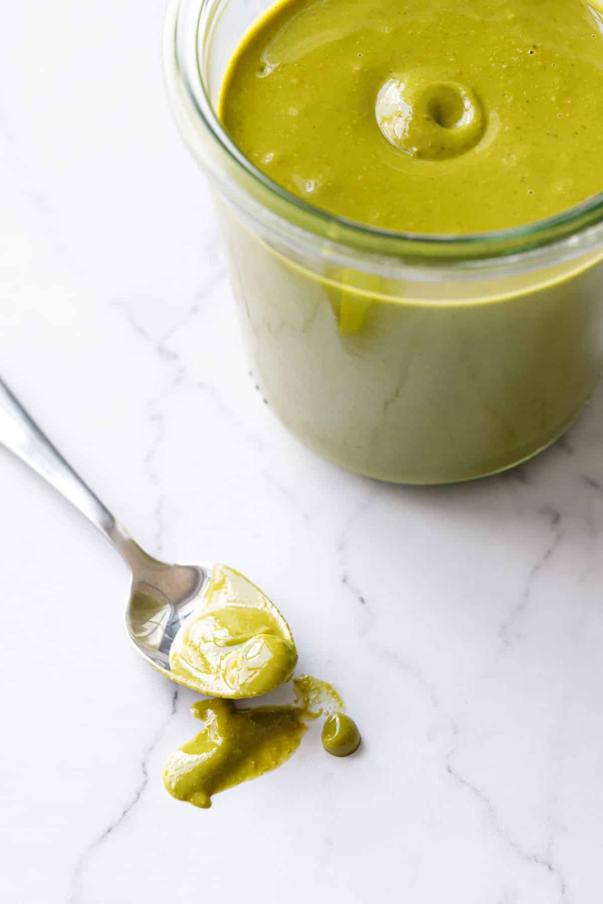 Spoonful of green homemade pistachio butter oozing onto a white marble background, with a jar of pistachio butter beside it.