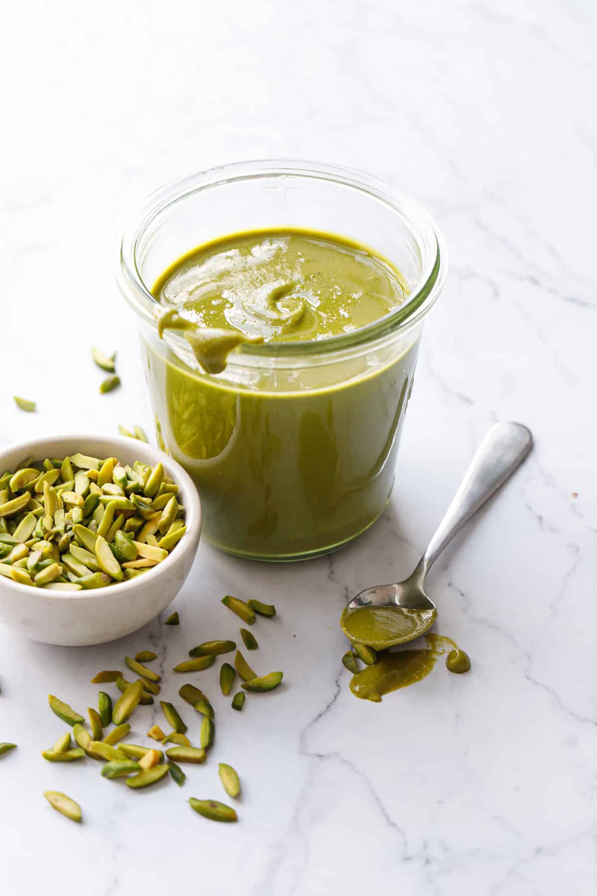 Jar of Homemade Pistachio Butter with a spoon oozing onto the table, with a bowl of slivered pistachios on the side and scattered around.