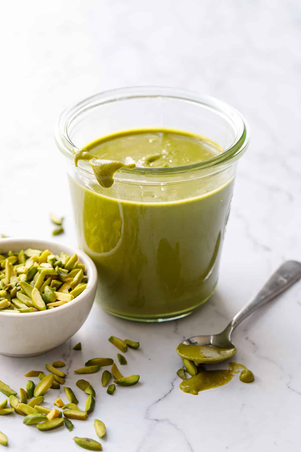 Jar of Homemade Pistachio Butter with a spoon oozing onto the table, with a bowl of slivered pistachios on the side and scattered around.