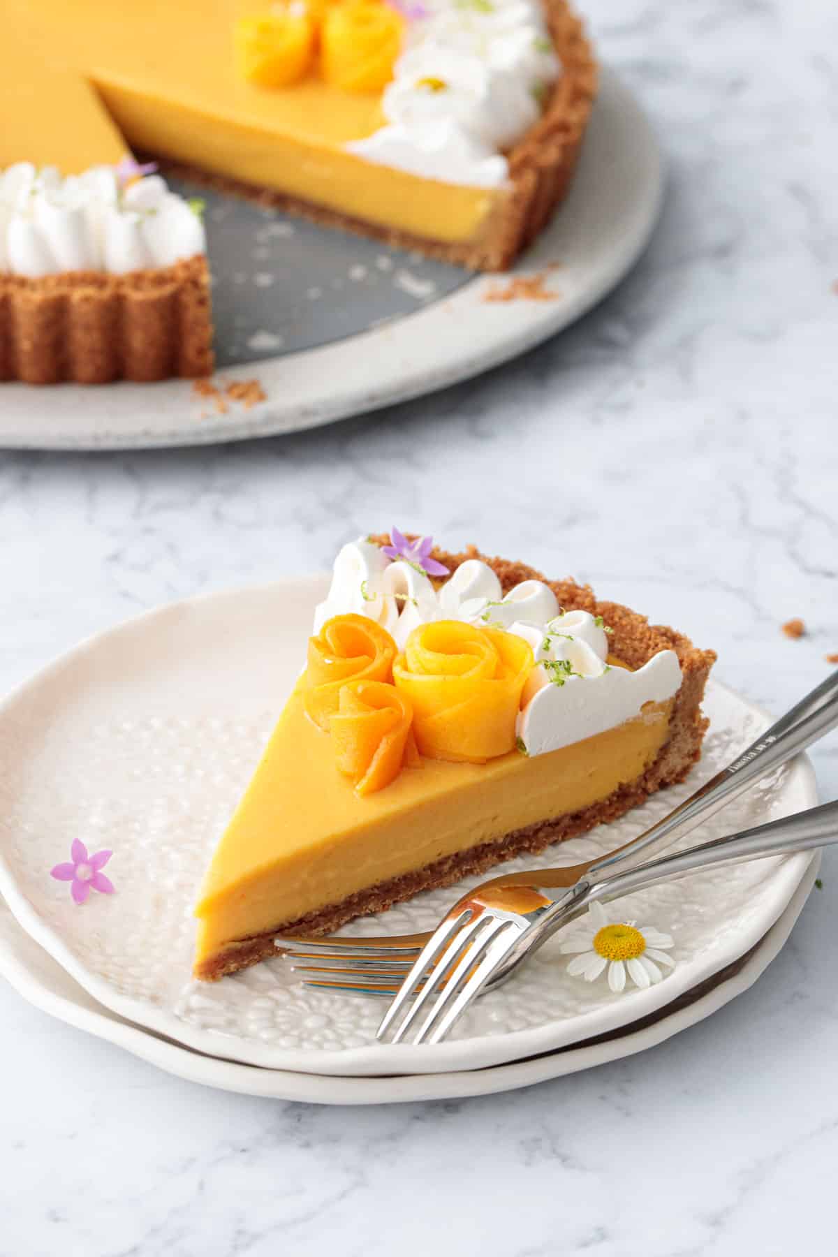 Cut slice of Mango Lime Tart on a white plate with two forks, topped with edible flowers and the rest of the tart in the background.